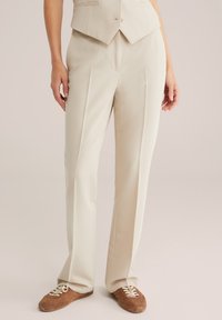 Beige skräddarsydda byxor med framfållar och en avslappnad passform, tillverkade av mjukt tyg, ihop med bruna snörskor.