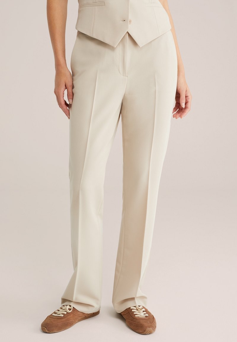 Beige skräddarsydda byxor med framfållar och en avslappnad passform, tillverkade av mjukt tyg, ihop med bruna snörskor.