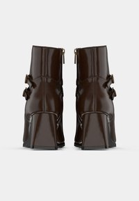 Botas de tobillo de charol marrón con un alto tacón cuadrado, que presentan dobles hebillas laterales y cierres de cremallera en la parte trasera. Textura lisa y brillante.