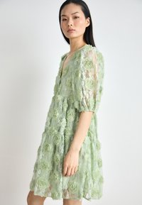 Robe en dentelle verte avec des appliqués floraux texturés, manches courtes bouffantes et décolleté en V. La jupe s'évasent à partir de la taille, créant du volume.