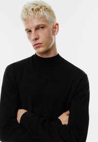 Svart polotröja i en slät stickad textur, figurnära design med långa ärmar, bärs av en person med korsade armar och kort hår.