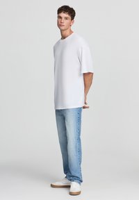 Maglietta bianca oversize con maniche corte, abbinata a jeans denim blu chiaro e sneakers bianche, in piedi su uno sfondo neutro.