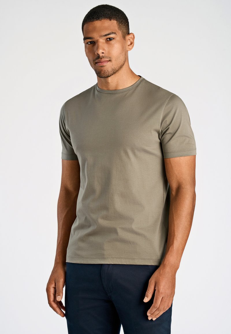 Mężczyzna o krótkich włosach, ubrany w oliwkowo-zieloną krótką koszulkę i ciemne spodnie, stojący na jasnym, jednolitym tle.