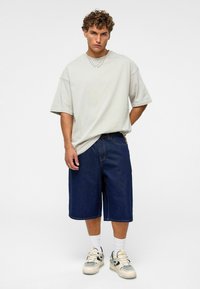 T-shirt oversize gris clair, shorts en denim foncé jusqu'aux genoux, chaussettes blanches et baskets beiges avec des accents noirs. Tenue simple et décontractée.