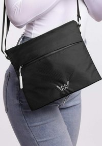 Černá crossbody taška z texturovaného materiálu, se zipovou kapsou vpředu a bílým logem jako akcentem. Nastavitelný popruh pro přizpůsobenou délku.