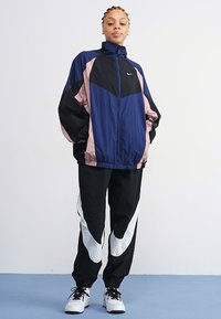 Giacca da track in navy, rosa e nero, con colletto alto e chiusura con zip. Abbinata a pantaloni neri con accenti bianchi sui lati e polsini elastici.