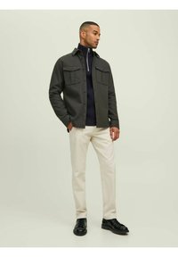 Camisa chaqueta de botón gris oscuro con dos bolsillos en el pecho, llevada sobre un suéter azul marino, combinada con pantalones crema y zapatos negros.