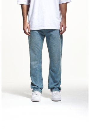 Homme portant un jean bleu clair ample, un t-shirt blanc et des baskets blanches, debout sur un fond blanc.
