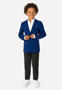 Zils bleizers ar atlokiem, divām priekšējām kabatām un izšuvumiem; balta krekls, melnas bikses un baltas kedas. Smaidošs bērns stāv pārliecinoši.