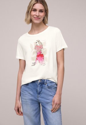 Smilende kvinde med kort blondt hår bærer en hvid T-shirt med en illustration af en kvinde i en rødstribet top og lyserødt skørt med teksten "Sommeren er min sæson."