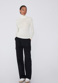 Langärmliger, enganliegender Rollkragenpullover in einem hellen Creme-Ton, kombiniert mit schwarzen Cargo-Hosen mit seitlichen Taschen, vervollständigt mit weißen Sneakers.