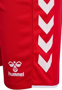 Czerwone sportowe spodenki z białymi paskami w kształcie jodełki po bokach oraz białym logo pszczoły Hummel w pobliżu brzegu.