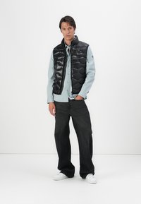 Gilet nero imbottito con finitura lucida, chiusura a zip e cuciture a forma di diamante, indossato sopra una camicia azzurra chiara, abbinato a jeans scuri e sneakers bianche.