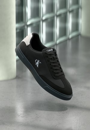 Melna zema augstuma Calvin Klein sporta apavu ar baltu "cK" logotipu sānos un gaiši pelēku papēža cilpu, redzami pelēka spoguļ virsmā lidojoši.