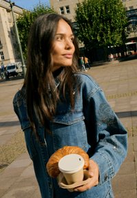 Femme en veste en jean tenant une tasse de café et un croissant en plein air sur une place urbaine avec des arbres et des bâtiments en arrière-plan.