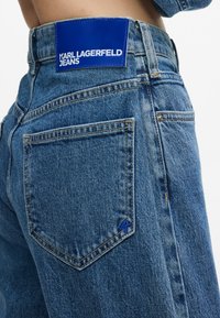 Gaiši zili džinsa bikses ar zilu etiķeti, uz kuras ir "KARL LAGERFELD JEANS." Ietver akcentu šuves un atpakaļsoma ar paraksta logo.