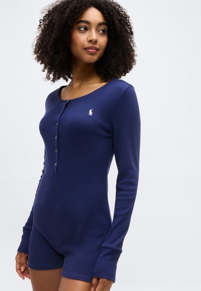 Γυναίκα με σγουρά μαλλιά που φοράει ένα σκούρο μπλε ολόσωμο σορτς με μακριά μανίκια, κουμπιά μπροστά και λευκό κεντημένο λογότυπο στο στήθος.