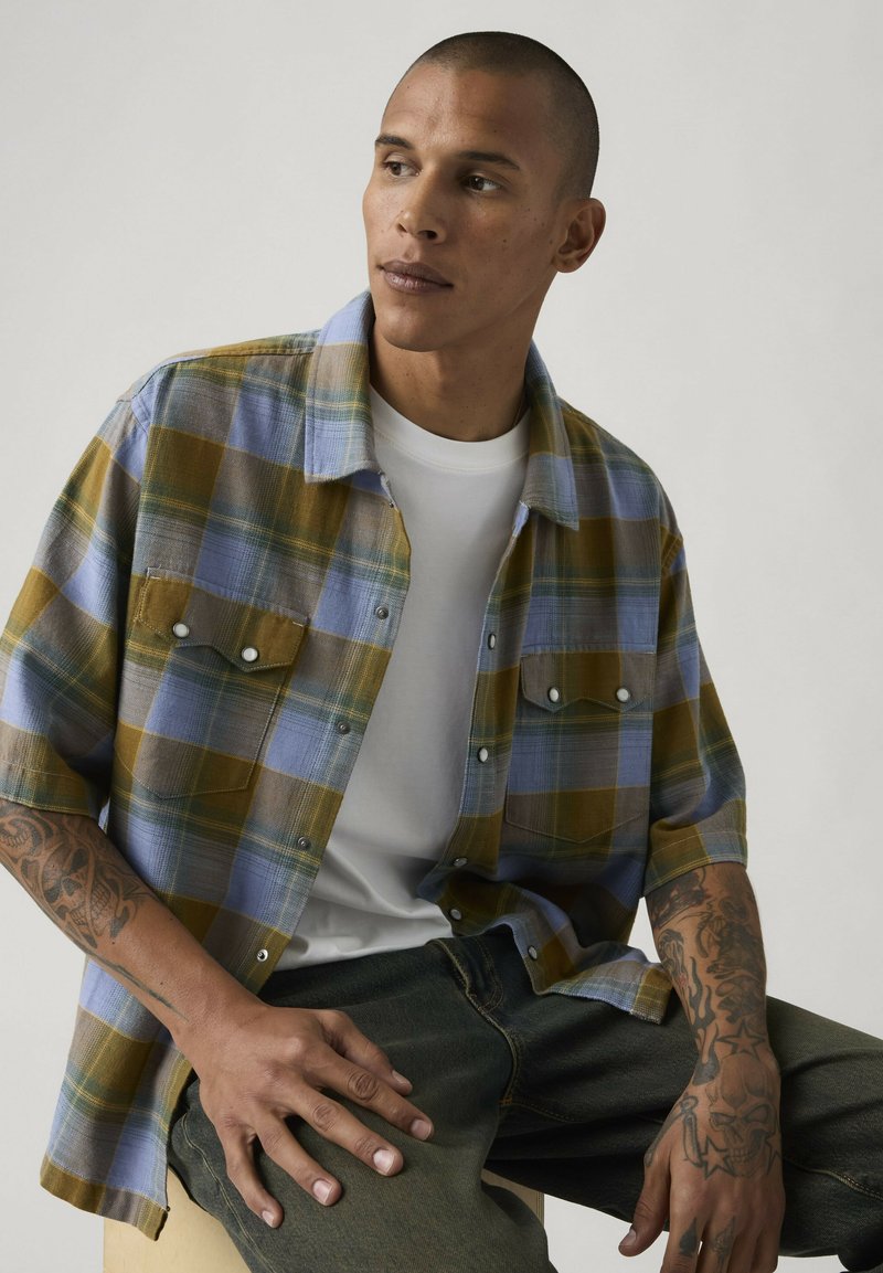 Man met tatoeages op beide armen, gekleed in een blauw-geel geruit overhemd over een wit T-shirt, zittend en zijwaarts kijkend.