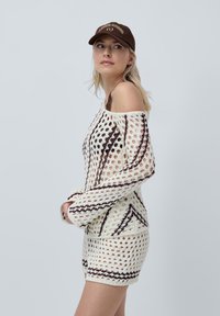 Abito in crochet crema con motivi geometrici marroni, design scoperto sulle spalle, maniche lunghe e silhouette aderente. Abbinato a un cappello marrone.