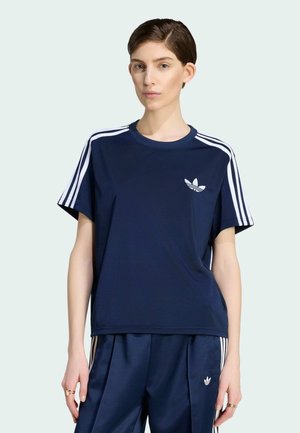 Kobieta w granatowym dresie Adidas z białymi paskami i logo, stoi na prostym jasnym tle.