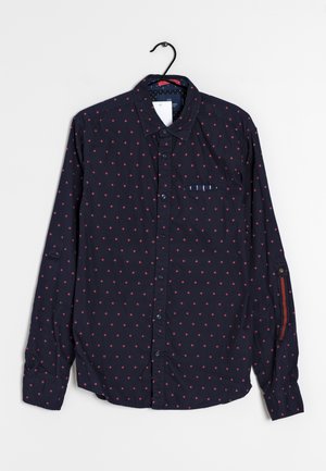 Chemise bleu marine à manches longues avec petit motif losange rouge, poche avant avec détail rayé, accrochée sur un cintre noir.