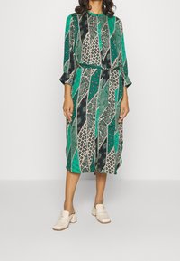 Grön mönstrad klänning med långa ärmar, prydd med olika geometriska och paisleymönster. Bärs med gräddfärgade träskor. Bälte vid midjan.