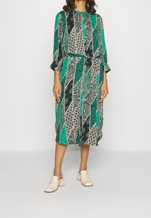 Vihreä kuvioitu mekko, jossa on pitkät hihat, ja se sisältää erilaisia geometrisia ja paisleymuotoja. Yhdistetty kermanvärisiin puukenkään. Vyötetty vyötäröltä.