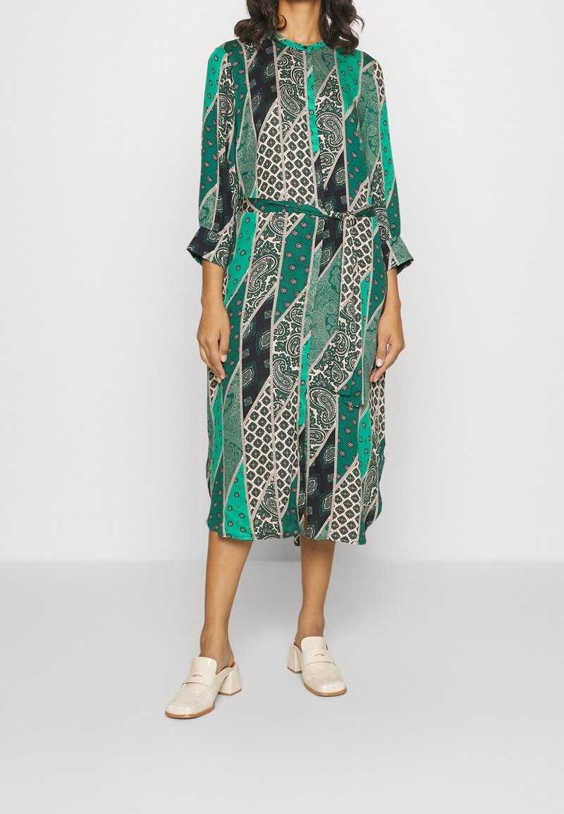 Zelené šaty s vzorem a dlouhými rukávy, které mají různé geometrické a paisley vzory. Kombinované s krémovými dřeváky. S páskem v pase.