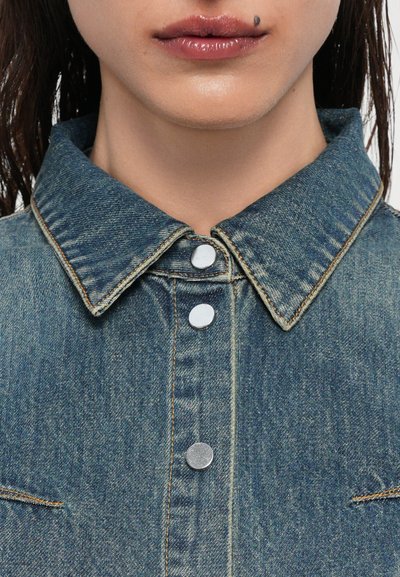 Camicia in denim con colletto classico, bottoni a pressione argentati e cuciture visibili in un blu medio con una texture liscia.