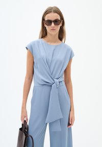 Tuta blu chiaro con maniche corte, tessuto annodato in vita e una texture fluida. Borsa nera tenuta in una mano.