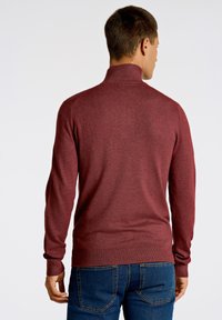 Maglione bordeaux con colletto alto, maniche lunghe e polsini a coste, realizzato in un morbido tessuto a maglia. Indossato con jeans in denim blu.