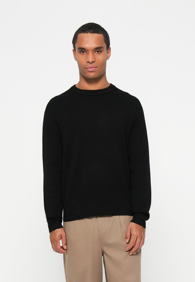 Sweater de gola redonda preto em um tecido macio, com mangas longas e um corte relaxado. A barra é ligeiramente mais longa na parte de trás.