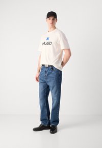 White cotton t-shirt with blue star and "HUGO" logo, paired with blue denim jeans and black shoes. The look is relaxed and casual.
