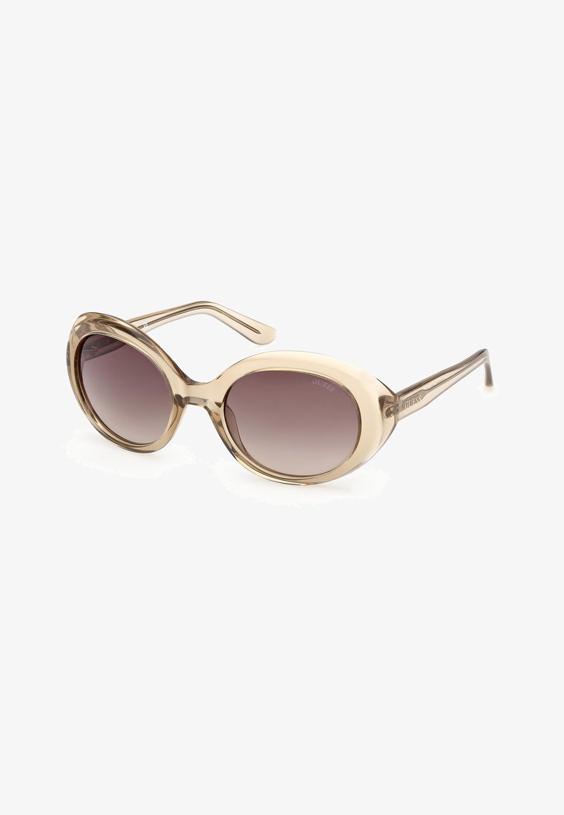 Lunettes de soleil ovales avec montures beiges translucides et verres dégradés marron foncé, logo GUESS visible sur les branches et les verres.