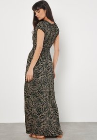 Maxikleid mit kurzen Ärmeln, schwarzem Grund, grünem und goldenem Blätter-Muster, geschopptem Bund, fließendem Rock und getragen mit flachen Sandalen.
