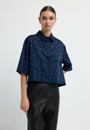 Donna che indossa una camicia azzurra a maniche corte con bottoni e piccole borchie argento, abbinata a pantaloni in pelle neri a vita alta, su sfondo bianco.