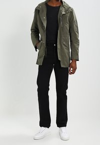 Parka vert olive avec capuche, fermeture éclair à l'avant et poches, portée sur une chemise noire et associée à un pantalon noir et des baskets blanches.