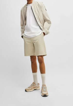 Homme portant une veste bomber beige, un t-shirt blanc, un short beige, des chaussettes blanches et des baskets beiges, debout les mains dans les poches.