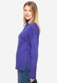 Lila Strickpullover mit hohem Kragen, gerippten Akzenten, seitlichen Schlitzen und langen Ärmeln; weicher Stoff und lockerer Schnitt. Kombiniert mit blauen Jeans.