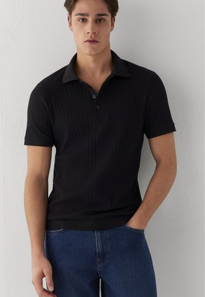 Junger Mann trägt ein schwarzes, kurzärmeliges, geripptes Poloshirt und blaue Jeans, steht vor einer weißen Wand mit einer Hand in der Tasche.