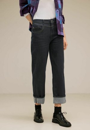 Persoon met opgerolde donkere jeans, zwarte veterschoenen, zwarte sokken, wit overhemd en paars-blauw geruit jasje, staand op een beige vloer.