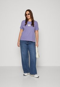 Purple cotton t-shirt with a circular graphic design at the chest, paired with wide-leg blue denim jeans and white sneakers.