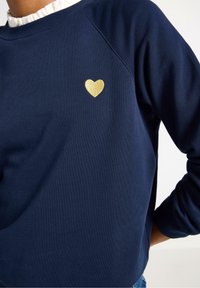 Tmavě modrý svetr s žebrovanou strukturou, raglánovými rukávy a zlatým glitrovým srdíčkem na hrudi. Pohodlný a ležérní design.