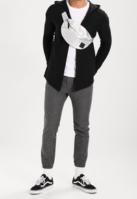 Sölj nylon midjeväska bärs över en svart cardigan, ihop med grårandiga joggers och svarta sneakers med vita sulor och strumpor.