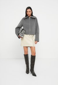 Γκρι κοντό σακάκι με φαρδιά μανίκια και ψηλό γιακά, συνδυασμένο με κρέμ δερμάτινη φούστα με φερμουάρ στο πλάι και μαύρες μπότες μέχρι το γόνατο.