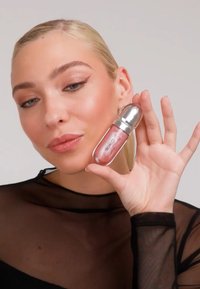 Femme aux cheveux blonds lisses et maquillage discret tenant un gloss à lèvres rose KIKO près de son visage, portant un haut noir transparent sur un fond uni.