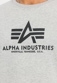 Hallide hall  rõivas, millel on must trükitud logo, geomeetrilised kujundid ja tekst "ALPHA INDUSTRIES" koos "KNOXVILLE, TENNESSEE, Ameerika Ühendriigid."