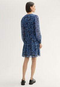 Bla dresses mit Blumenmuster aus transparentem Stoff, an der Taille gerafft, mit langen Ärmeln und einem gestuften Saum. Schwarze Schuhe vervollständigen das Outfit.