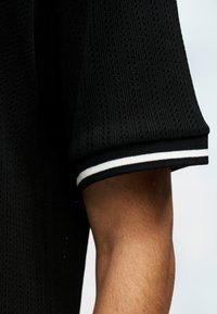 Μαύρο πλεκτό πουκάμισο με κοντό μανίκι και διπλές λευκές ρίγες στη μανσέτα, φορεμένο σε χέρι μπροστά σε μονόχρωμο φόντο.