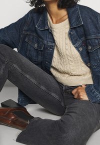 Veste en denim avec poches, pull léger à torsades, jean gris foncé et bottines en cuir marron avec une surface texturée.